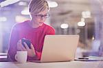 Serious businesswoman working late, using cell phone at laptop in office