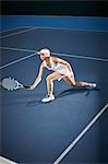 Young female tennis player playing tennis, reaching with tennis racket on blue tennis court