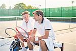 Young male tennis players resting with tennis rackets on sunny tennis court