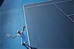 Overhead view young male tennis player playing tennis, serving the ball on sunny blue tennis court