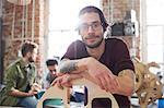 Portrait confident male designer with tattoos leaning on prototype in workshop