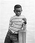 1970s BAHAMIAN BOY IN STRIPED SHIRT WITH SAD FACIAL EXPRESSION LOOKING AT CAMERA ON DOCK HOLDING UP TWO VERY SMALL FISH