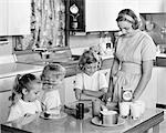 1950s MOTHER & THREE DAUGHTERS STANDING AT KITCHEN TABLE MAKING LUNCH PEANUT BUTTER & JELLY SANDWICHES & GLASSES OF MILK