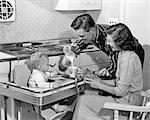 1950s FATHER POURING MILK INTO CUP FOR SON IN HIGH CHAIR WITH MOTHER SEATED AT SIDE