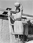 1930s NATIVE AMERICAN INDIAN WOMAN STANDING ON LADDER HOLDING OLLA WATER JAR POTTERY ON HER HEAD COCHITI PUEBLO NEW MEXICO USA