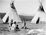 1920s NATIVE AMERICAN INDIAN FAMILY MAN WOMAN TWO CHILDREN SITTING IN FRONT OF TEPEES SIOUX TRIBE MONTANA USA