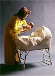 1970s MOTHER WEARING YELLOW ROBE PUTTING BABY CHILD TO SLEEP IN WHITE WOVEN WICKER BASSINET