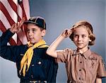 1960s BOY AND GIRL IN CUB SCOUT AND BROWNIE UNIFORMS STANDING BY AMERICAN FLAG SALUTING