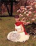 1960s YOUNG WOMAN SITTING BY BLOSSOMING MAGNOLIA TREE WITH SPRINGTIME HAT RED SCARF WHITE GLOVES AND DRESS LOOKING AT CAMERA