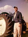 1960s PORTRAIT OF SERIOUS MAN FARMER STANDING BESIDE LARGE TRACTOR TIRE LOOKING AT CAMERA