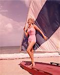 1960s BLOND WOMAN BEAUTY IN PINK SWIM WEAR BATHING SUIT POSED ON BLUE WHITE SAIL BOAT SAILFISH BOAT ON SUMMER SEASHORE BEACH