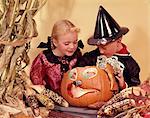 1960s TWO KIDS BLOND BROTHER AND SISTER WEARING HALLOWEEN COSTUMES LOOKING INTO CARVED JACK-O-LANTERN PUMPKIN