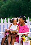 1990s MOTHER SITTING ON WHITE LAWN CHAIR WITH DAUGHTER ON HER LAP WITH BOOK