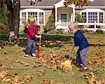 1960s BOY GIRL RAKING AUTUMN LEAVES IN YARD SUBURBAN HOUSE IN BACKGROUND