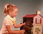 1960s GIRL WITH BLONDE PONYTAIL PLAYING WITH TOY DOLLHOUSE