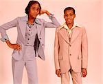 1970s TWO AFRICAN AMERICAN TEENAGERS A BOY IN A BEIGE SUIT AND GIRL IN A BLUE PANTS SUIT STANDING TOGETHER LOOKING AT CAMERA