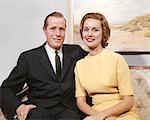 1960s HAPPY COUPLE SITTING ON SOFA MAN WEARING BUSINESS SUIT TIE WOMAN IN A YELLOW DRESS LOOKING AT CAMERA SMILING