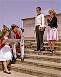 1960s 4 TEENS ON FRONT STEPS HIGH SCHOOL BUILDING TALKING HOLDING BOOKS