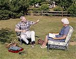 1960s OLDER COUPLE SITTING IN YARD WOMAN READING MAN WORKING WITH FISHING TACKLE GEAR