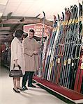 1960s COUPLE LOOKING AT SKIS IN SPORTS DEPARTMENT STORE LIFESTYLE VACATION MAN WOMAN WEARING COATS
