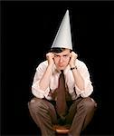 1960s UNHAPPY YOUNG MAN SITTING ON STOOL WEARING DUNCE CAP LOOKING AT CAMERA