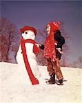 1970s LAUGHING BOY BUILDING SNOWMAN BOTH WEARING RED KNIT SCARVES IN COLD WINTER SNOW