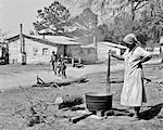 1930s ELDERLY MAN AND GRANDCHILDREN WALKING BY AFRICAN AMERICAN WOMAN STIRRING CALDRON ON OPEN FIRE ON TURPENTINE PLANTATION