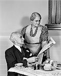 1940s ELDERLY COUPLE MAN WOMAN IN KITCHEN MAN SITTING AT TABLE WOMAN LOOKING OVER HIS SHOULDER READING NEWSPAPER