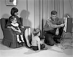 1960s FAMILY OF FOUR WATCHING PORTABLE TELEVISION MOTHER DAUGHTER IN CHAIR FATHER TURNING TV CHANNEL DIAL SON SITTING ON RUG