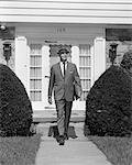 1950s BUSINESSMAN LEAVING SUBURBAN HOUSE HOLDING BRIEFCASE WALKING DOWN SIDEWALK