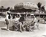 1930s 1940s 3 WOMEN BATHING SUITS SINGLE MAN CASUAL CLOTHES SITTING TALKING UNDER POOL SIDE UMBRELLA MIAMI BEACH FLORIDA USA