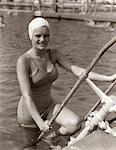 1930s SMILING YOUNG WOMAN WEARING ONE PIECE BATHING SUIT & CAP CLIMBING OUT OF POOL LOOKING AT CAMERA ON VACATION IN FLORIDA USA