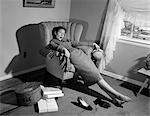 1950s WOMAN EXHAUSTED FROM SHOPPING SLUMPING IN LIVING ROOM CHAIR SHOES OFF PACKAGES EVERYWHERE LOOKING AT CAMERA