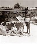 1930s 1940s MAN BATHING TRUNKS TALKING TO TWO WOMEN LAYING UNDER HOTEL CANVAS SUN SCREEN TROPICAL SAND MIAMI BEACH FLORIDA USA