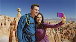 Boyfriend and girlfriend taking selfie with smartphone near scenic rock formations in remote landscape under clear blue sky