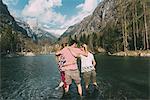 Rear view of three young adult friends ankle deep in mountain lake, Lombardy, Italy