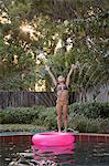 Young girl standing on edge of outdoor swimming pool, in front of sprinkler, arms raised