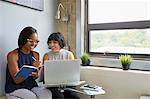 Woman working on laptop, on laptop stand, colleague sitting beside her writing in notebook