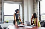 Woman sitting at desk with laptop, talking to female colleague standing beside desk