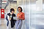 Two businesswomen standing in office corridor, looking at digital tablet, smiling