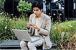 Young man sitting outdoors, using laptop, holding smartphone