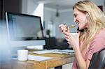 Woman at computer making telephone call on smartphone while eating breakfast