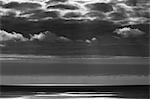 View from the land over the ocean, to the horizon in low light. Sunlight patches on the water surface and dark storm clouds overhead.