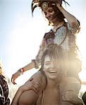 Young woman at a summer music festival carrying a friend on her shoulders, face painted, smiling at camera.