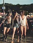 Three young women at a summer music festival wearing hot pants and Wellington boots, feather headdress and faces painted, arms around shoulder and smiling.
