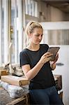 Woman with blond hair standing in a workshop using a digital tablet, wrapped parcels on a table.