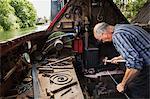 Blacksmith working in a small space on his narrowboat, a barge on river, bending over the anvil and shaping hot metal.