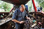 Blacksmith standing in his workshop, a floating forge on a narrowboat barge, working on a metal object, using a file.