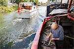 High angle view of a blacksmith working at his bench on a narrowboat on the water, holding a tape measure. A motorboat underway.