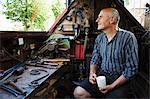 Blacksmith sitting on a working narrowboat on a waterway, in his workshop holding mug.